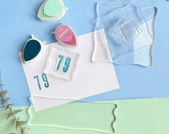 Puede incluir: Un conjunto de bloques de sellos de acrílico transparente con líneas de cuadrícula, dos almohadillas de tinta en forma de gota en verde menta y rosa, y un sello transparente pequeño con el número 79, con un 79 azul turquesa estampado en una tarjeta blanca.