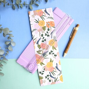 Puede incluir: Una guillotina de papel de color morado claro con una gu&iacute;a de corte de pl&aacute;stico transparente y una tarjeta rectangular blanca con un estampado floral de flores naranjas, amarillas y rosas con hojas verdes y peque&ntilde;os p&aacute;jaros azules, junto a un bol&iacute;grafo de madera.