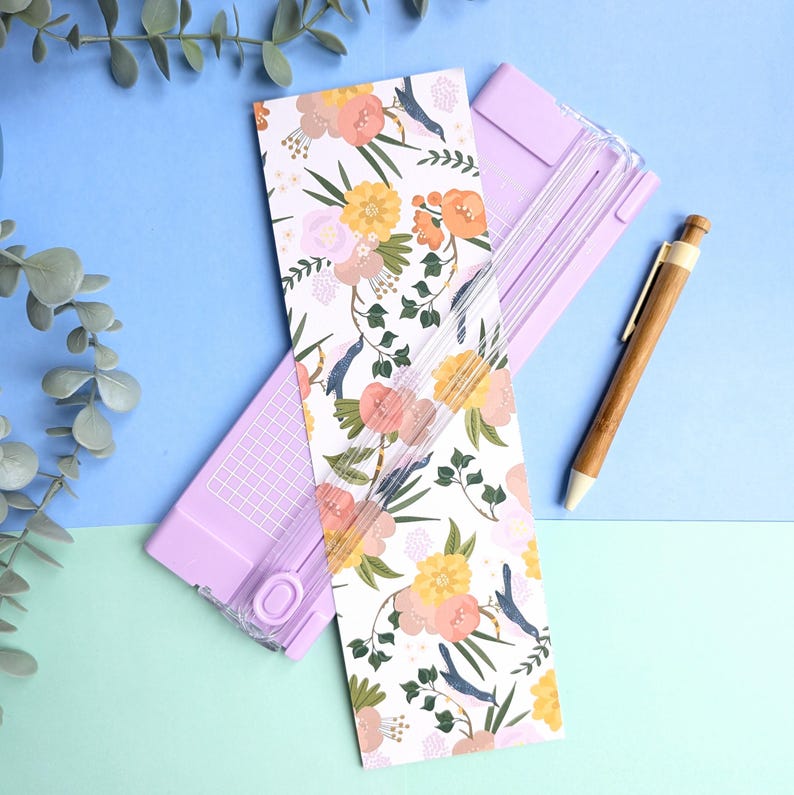 Puede incluir: Una guillotina de papel de color morado claro con una gu&iacute;a de corte de pl&aacute;stico transparente y una tarjeta rectangular blanca con un estampado floral de flores naranjas, amarillas y rosas con hojas verdes y peque&ntilde;os p&aacute;jaros azules, junto a un bol&iacute;grafo de madera.