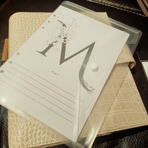 May include: A beige planner with a textured, crocodile-like pattern. A clear plastic envelope with a white button closure sits on top, holding a lined paper with the letter "M" and the word "MAY" printed on it. A pen is visible.