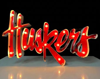 May include: A red marquee-style sign featuring the word Huskers in a cursive script font. The sign is illuminated by numerous small, warm-toned light bulbs embedded along the surface of the letters, creating a bright and glowing display.