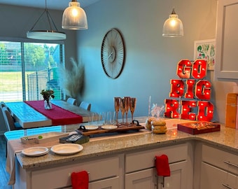 May include: A white kitchen island with a granite countertop and white cabinets. The island has a red towel hanging from a cabinet handle. There are glasses and a tray of snacks on the counter. The wall behind the island is painted a light blue color and has a red light-up sign that says "Go Big Red".