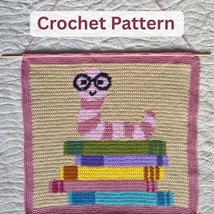 May include: A crocheted wall hanging with a pink and white striped worm wearing glasses, sitting on a stack of colourful books. The background is beige, and the border is pink. The text "Crochet Pattern" is at the top.