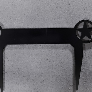 May include: Black metal decorative piece featuring a star within a circle on each end, connected by a horizontal bar. The design includes two downward-pointing legs. The star is a five-pointed star.