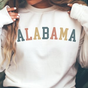 May include: White sweatshirt with the word "ALABAMA" printed in rainbow colors across the front.