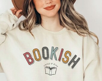 Può includere: Una felpa color crema con la parola "BOOKISH" in colori arcobaleno e una grafica stilizzata di un libro aperto. La modella indossa un cappello marrone e jeans blu.