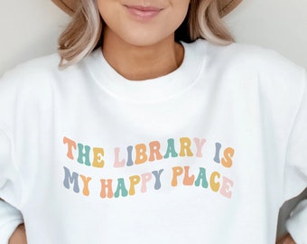 Peut inclure: Sweat-shirt blanc avec l'inscription "THE LIBRARY IS MY HAPPY PLACE" en lettres colorées de style rétro. Le haut à manches longues est de style décontracté, adapté au quotidien.