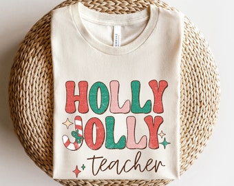 May include: A beige t-shirt with the text "HOLLY JOLLY teacher" in red, green, and pink letters with a candy cane and stars.