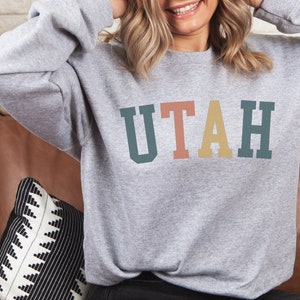 May include: A grey sweatshirt with the word "UTAH" printed in colourful block letters. The letters are in shades of teal, pink, yellow, and green.