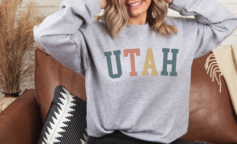 May include: A grey sweatshirt with the word "UTAH" printed in colourful block letters. The letters are in shades of teal, pink, yellow, and green.