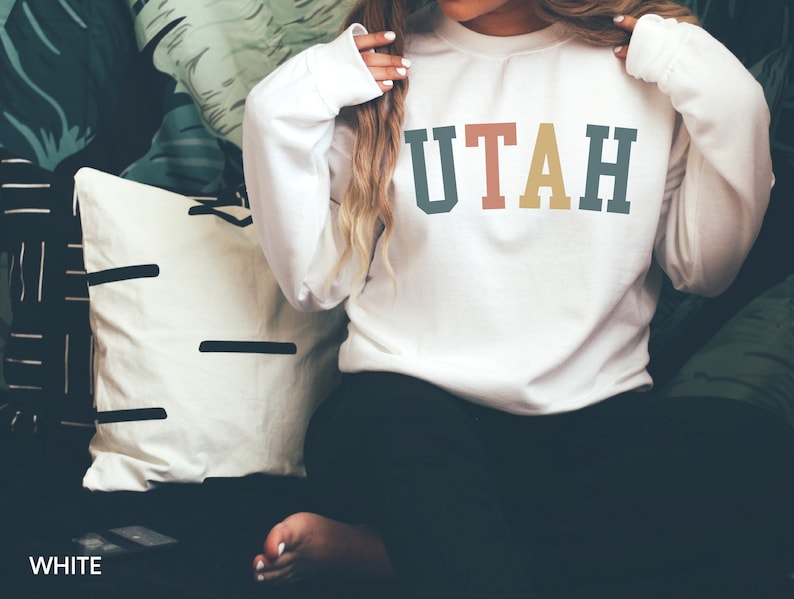 May include: White sweatshirt with the word "UTAH" printed in colourful block letters. The sweatshirt is being worn by a person sitting on a sofa.