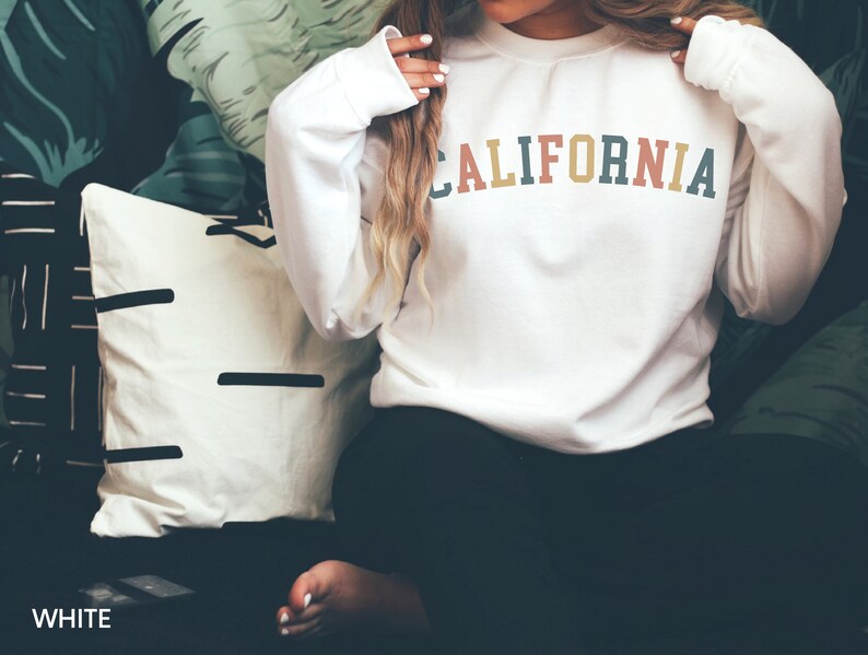 Puede incluir: Sudadera blanca con un estampado de "CALIFORNIA" de colores en la parte delantera.