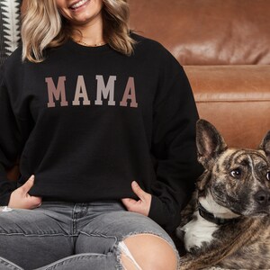 May include: A woman wearing a black sweatshirt with the word "MAMA" printed in a gradient of pink and brown. She is sitting on a brown leather couch with a dog beside her.