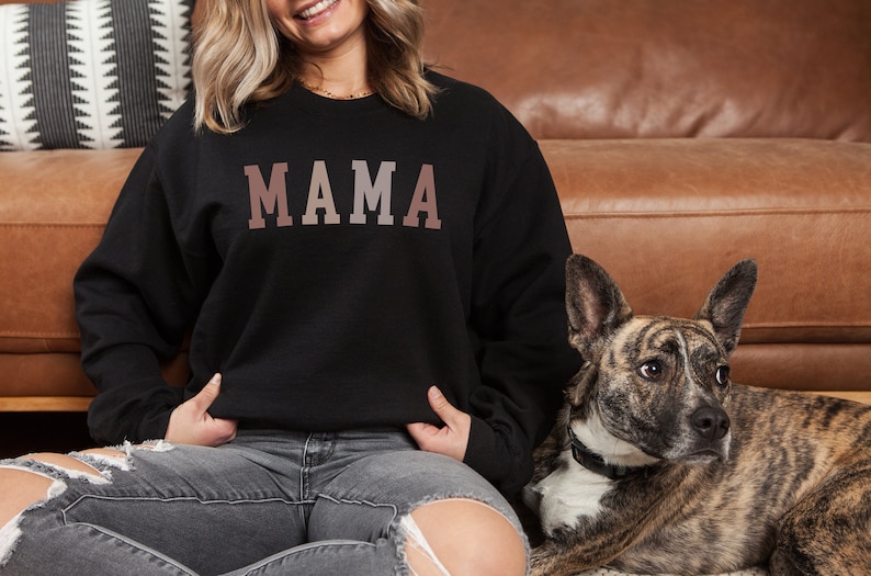 May include: A woman wearing a black sweatshirt with the word "MAMA" printed in a gradient of pink and brown. She is sitting on a brown leather couch with a dog beside her.