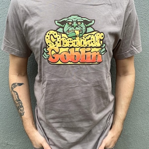 Könnte beinhalten: Ein heidefarbenes T-Shirt mit einem Aufdruck eines grünen Kobolds mit dem Text "Cheddar Goblin" in Gelb, Orange und Rot.