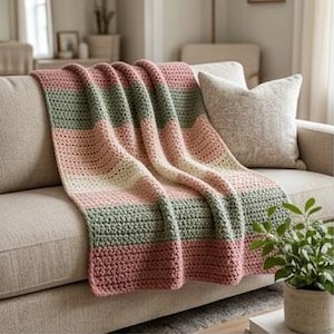 Puede incluir: Una manta de ganchillo rectangular con rayas horizontales en tonos alternos de rosa empolvado, verde salvia y crema. El tejido presenta un patrón de puntos texturizado y consistente, drapeado sobre un sofá de color neutro.