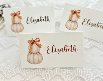 May include: Three white rectangular place cards featuring a watercolor illustration of two stacked pumpkins topped with an orange ribbon bow. The name Elizabeth is printed in a dark script font on each card. The cards are arranged in a group.