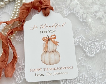May include: A white rectangular gift tag with rounded corners and a scalloped top edge, featuring a watercolor illustration of two stacked pumpkins with an orange ribbon bow. The text reads: So thankful FOR YOU, HAPPY THANKSGIVING! Love, The Johnsons.