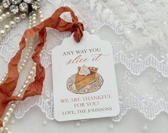 May include: A white rectangular gift tag with scalloped edges, featuring a watercolor illustration of a pumpkin pie slice on a floral plate. The text reads: ANY WAY YOU slice it, WE ARE THANKFUL FOR YOU!, LOVE, THE JOHNSONS. Attached with a rust-colored ribbon.