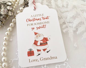 May include: A white gift tag with red and white twine. The tag features a cartoon illustration of Santa Claus holding a gift box. The text on the tag reads "A little Christmas treat for someone so sweet! Love, Grandma".