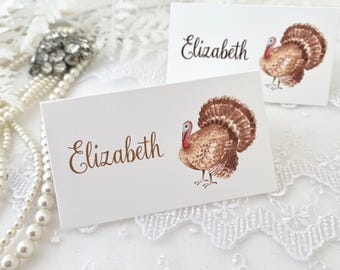 May include: Two white place cards with the name "Elizabeth" written in brown cursive. Each card features a watercolor illustration of a brown turkey with a red head and a white background.