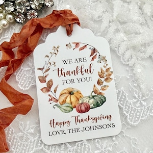 Könnte beinhalten: Ein weißes, rechteckiges Geschenkanhänger-Schild mit gewelltem Rand und einem Aquarellkranz aus Herbstblättern und Kürbissen. Der Text lautet WE ARE thankful FOR YOU! Happy Thanksgiving LOVE, THE JOHNSONS. Ein rostfarbenes Band ist befestigt.