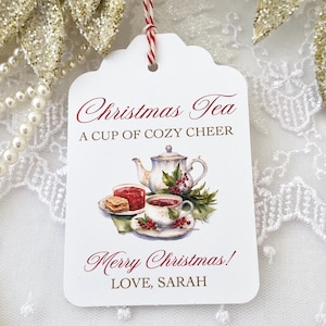 Könnte beinhalten: Ein weißes Geschenk-Etikett mit gewellten Kanten, mit dem Text "Christmas Tea" und "A Cup of Cozy Cheer". Das Etikett enthält eine Aquarell-Illustration einer Teekanne, einer Teetasse und Leckereien. Das Etikett ist mit rot-weißem Bindfaden gebunden.
