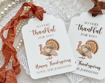 May include: Two white rectangular gift tags with rounded corners, each featuring a brown turkey illustration. The text reads SO VERY thankful FOR YOU! Happy Thanksgiving LOVE, THE JOHNSONS. Each tag has a rust-colored ribbon tied at the top.