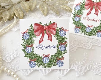 May include: A white card with a watercolor illustration of a green wreath with red bow and blue and white ornaments. The name "Elizabeth" is written in blue cursive on the card.