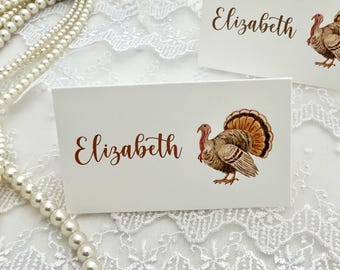 May include: A white rectangular place card featuring the name Elizabeth printed in a brown script font alongside a detailed illustration of a brown and gold turkey. The card is positioned on a white lace surface with a string of white pearls.