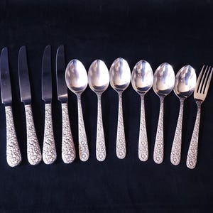May include: A set of silver cutlery, comprising knives, spoons, and a fork, displayed on a dark background. The handles of the knives and spoons are decorated with a floral design. The spoons have a rounded bowl.