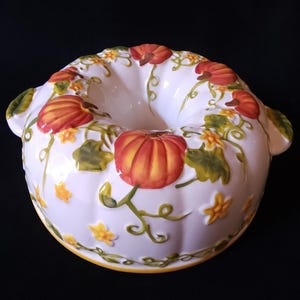 May include: White ceramic bundt pan with a pumpkin and vine design. The pan features orange and yellow pumpkins, green leaves, and yellow flowers. The rim has a yellow border and two handles. The design is hand-painted.