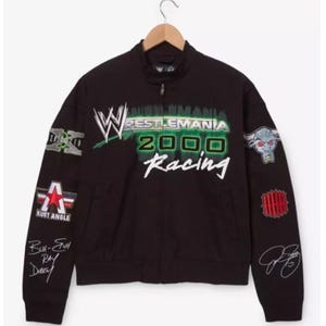 May include: A black zip-up jacket featuring a large WrestleMania 2000 Racing graphic on the chest. The sleeves are decorated with various patches, including a bull head, a red star with the text Kurt Angle, and several embroidered signatures.