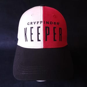 Puede incluir: Gorra de béisbol con diseño dividido, con paneles blancos y rojos. La visera y la corona negras se complementan con el texto bordado "GRYFFINDOR KEEPER" en negro, creando un contraste audaz. La gorra es un artículo para fans.