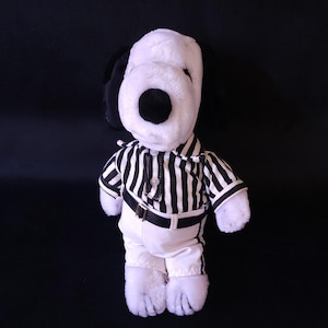 Könnte beinhalten: Ein Snoopy-Plüschtier, das ein schwarz-weiß gestreiftes Schiedsrichterhemd, eine weiße Hose und einen schwarzen Gürtel trägt. Das Spielzeug hat schwarze Ohren und eine schwarze Nase. Der Hintergrund ist einfarbig schwarz.