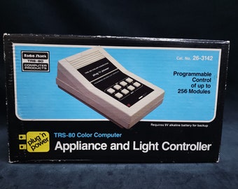 Op de afbeelding: Een witte Radio Shack TRS-80 Color Computer plug 'n power apparaat- en lichtcontroller doos. De doos heeft een afbeelding van het apparaat, dat een witte kunststof behuizing heeft met een zwart display en een rij knoppen. De tekst op de doos luidt "TRS-80 Color Computer Appliance and Light Controller" en "Programmable Control of up to 256 Modules".