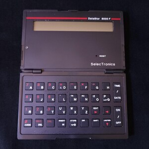 Puede incluir: Un organizador electrónico DataStor 8000 F negro y rojo vintage con un teclado completo y una pantalla. El dispositivo tiene un diseño con bisagras y presenta la marca "SelecTronics". El teclado tiene teclas rojas con letras blancas.