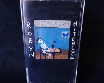 May include: A clear plastic cassette case featuring the album cover for The Man Upstairs by Robyn Hitchcock. The artwork depicts a skeleton wearing a hat, sitting at a table in a blue room with a blue butterfly and a framed butterfly picture.