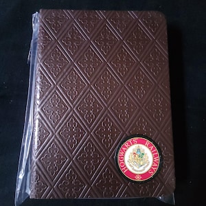 May include: A brown notebook with a diamond pattern and a Hogwarts Railways emblem. The emblem is a circular badge with a red border and the Hogwarts crest in the center. The notebook is in a clear plastic sleeve.