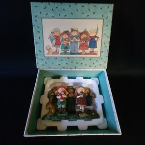 May include: A decorative box with a painted illustration of a group of children and a dog. Inside, a figurine set depicts children holding flowers and gifts. The box is teal with a white interior and a matching lid.