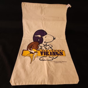 Puede incluir: Bolso tote color crema con Snoopy con un casco de fútbol morado sosteniendo un balón de fútbol. El logo de los Minnesota Vikings está debajo. El bolso tiene un cierre de cremallera en la parte superior.