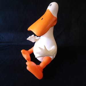 May include: A cartoon-style duck figurine featuring a smooth white body and a large, prominent orange beak. The duck has orange feet and hands, with simple black eyes positioned on the upper white head. It is depicted in a seated position.
