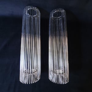 Może przedstawiać: Dwa cylindryczne, srebrne metalowe świeczniki. Każdy uchwyt składa się z pionowych metalowych prętów, tworząc otwartą konstrukcję przypominającą klatkę. Góra i dół są okrągłe, a pręty połączone tworzą konstrukcję. Tło jest jednolicie czarne.