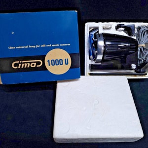 Puede incluir: Una lámpara universal negra Cima 1000 U para cámaras fijas y de cine en su caja original. La lámpara tiene una carcasa de metal negro con una perilla plateada y un cable negro. La caja es azul con letras blancas y un círculo dorado con el texto "Cima 1000 U".