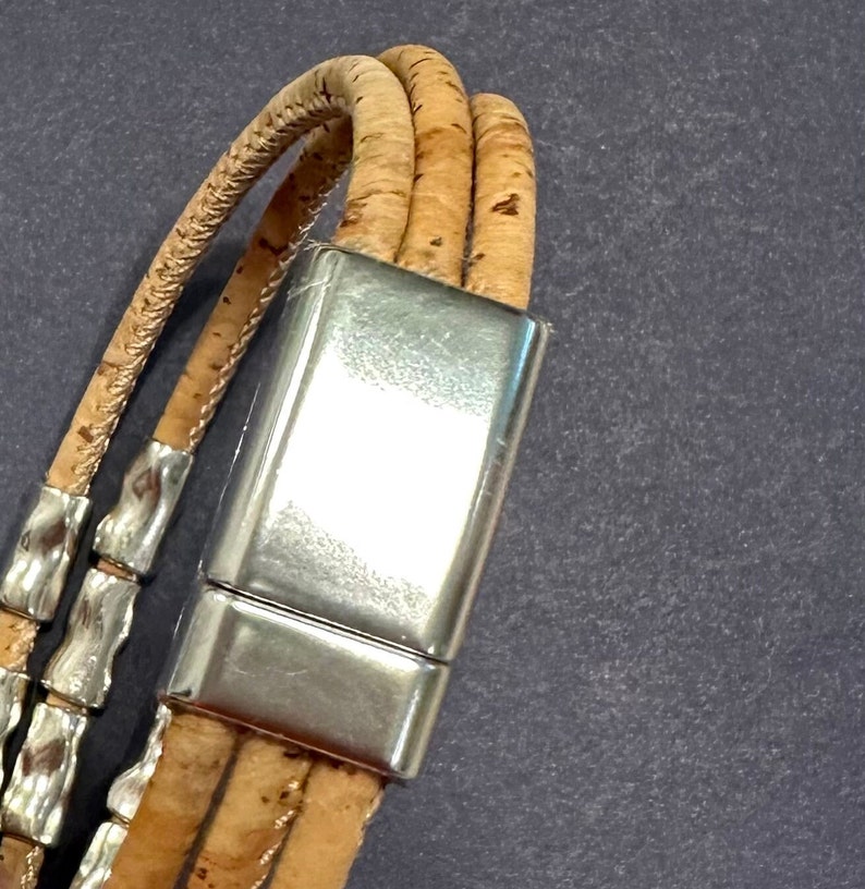 Op de afbeelding: Een armband met meerdere strengen gemaakt van natuurlijk bruin kurkkoord, voorzien van een gepolijste zilverkleurige rechthoekige magnetische sluiting en kleine, getextureerde zilverkleurige metalen cilindrische kralen.