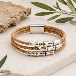 Op de afbeelding: Een meerstrengs armband gemaakt van natuurlijk bruin kurkkoord, voorzien van drie rijen gladde, gepolijste zilverkleurige metalen kralen en een rechthoekige zilverkleurige magnetische sluiting. Het ontwerp bestaat uit drie parallelle kurkstrengen.