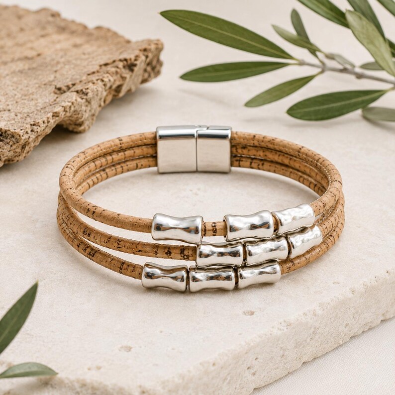 Op de afbeelding: Een meerstrengs armband gemaakt van natuurlijk bruin kurkkoord, voorzien van drie rijen gladde, gepolijste zilverkleurige metalen kralen en een rechthoekige zilverkleurige magnetische sluiting. Het ontwerp bestaat uit drie parallelle kurkstrengen.