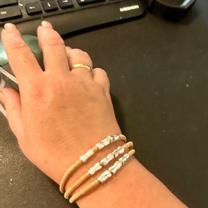 Op de afbeelding: Een armband met drie strengen gemaakt van lichtbruin kurkmateriaal. Elk van de drie strengen is voorzien van twee kleine, onregelmatige zilverkleurige metalen kralen die gelijkmatig over de lengte van het kurkkoord zijn verdeeld.