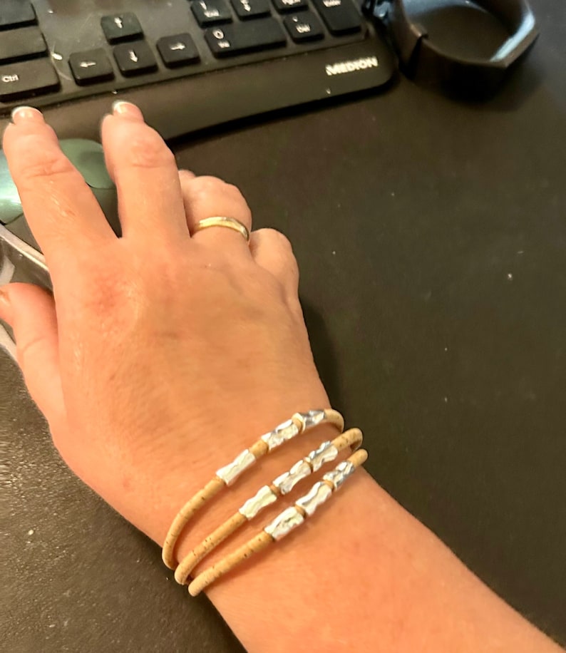 Op de afbeelding: Een armband met drie strengen gemaakt van lichtbruin kurkmateriaal. Elk van de drie strengen is voorzien van twee kleine, onregelmatige zilverkleurige metalen kralen die gelijkmatig over de lengte van het kurkkoord zijn verdeeld.