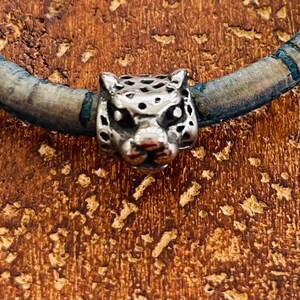 May include: A silver charm bracelet with a detailed silver leopard head charm. The bracelet is made of a light blue leather material.